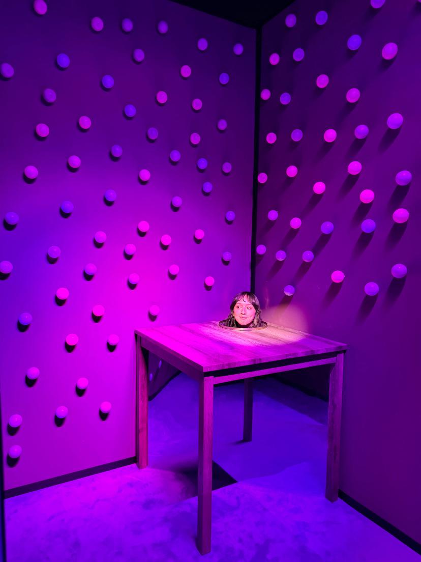 Interactive art installation featuring a woman’s head appearing through a wooden table in a room illuminated with purple and pink lighting and walls covered in circular designs