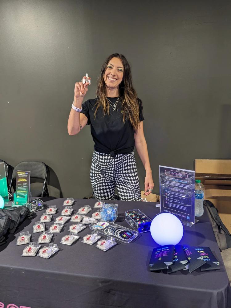 Smiling woman standing behind a booth displaying educational materials, promotional items, and merchandise for a 'Keep the Party Safe' campaign.