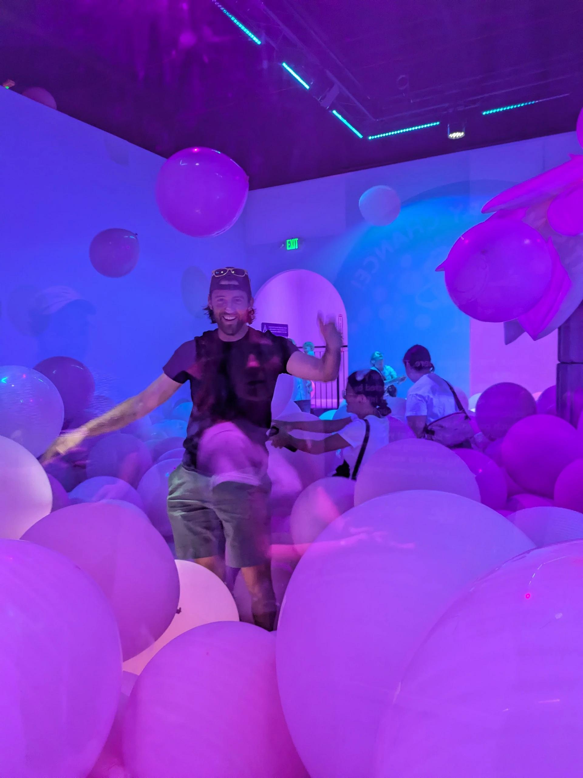 A man smiling and playing in a room filled with large pink and purple balloons under vibrant neon lighting, creating a playful and immersive atmosphere.